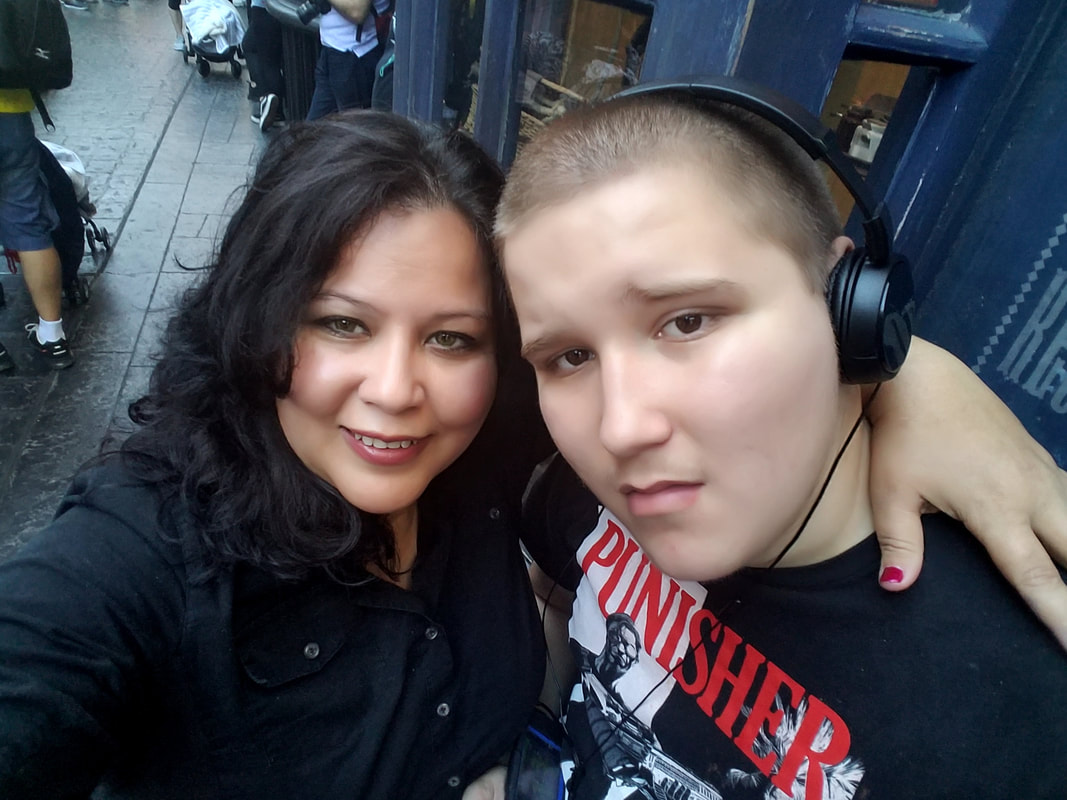Mother on the left and autistic son wearing headphones on the right. They are cheek to cheek posing for the picture. Mom is smiling and son looks pensive. Mom is wearing black blouse, son is wearing a Punisher black shirt.